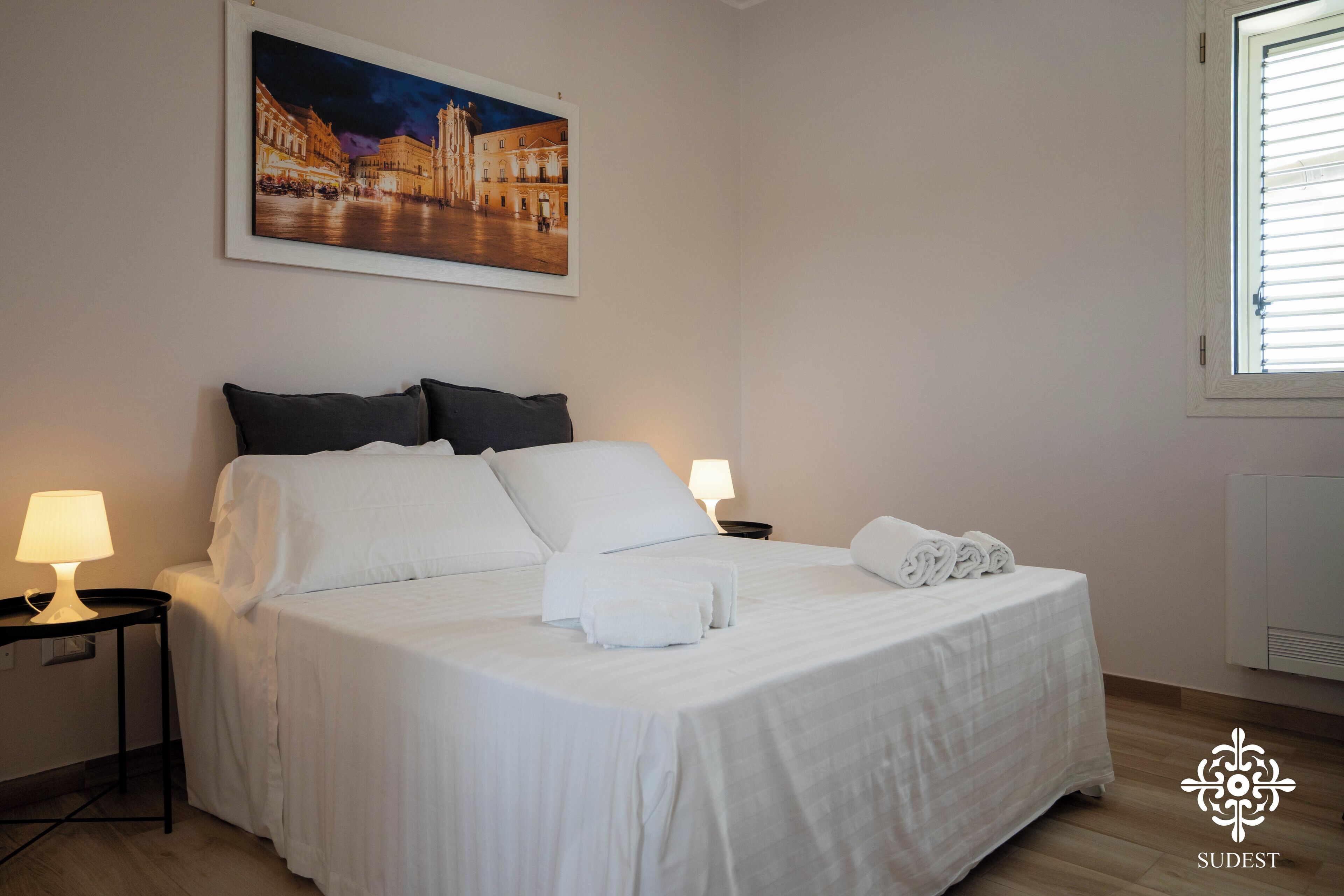 A neatly made bed with white linens, two bedside tables with lamps, a framed picture on the wall, and a window with blinds.