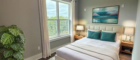 Une chambre avec un grand lit, deux tables de chevet avec des lampes, un tableau représentant un paysage, une fenêtre avec des rideaux et une plante en pot.