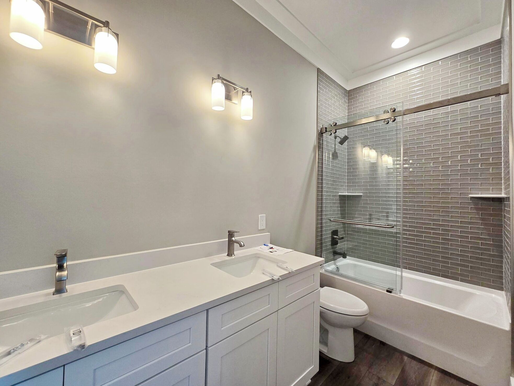 A modern bathroom with a double sink vanity, a toilet, and a glass-enclosed shower with a brick backsplash.