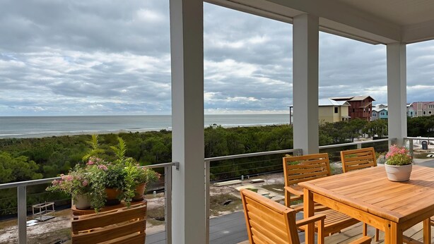 Un espacio al aire libre techado con una mesa y sillas de madera, plantas en macetas y vistas al océano y la playa.