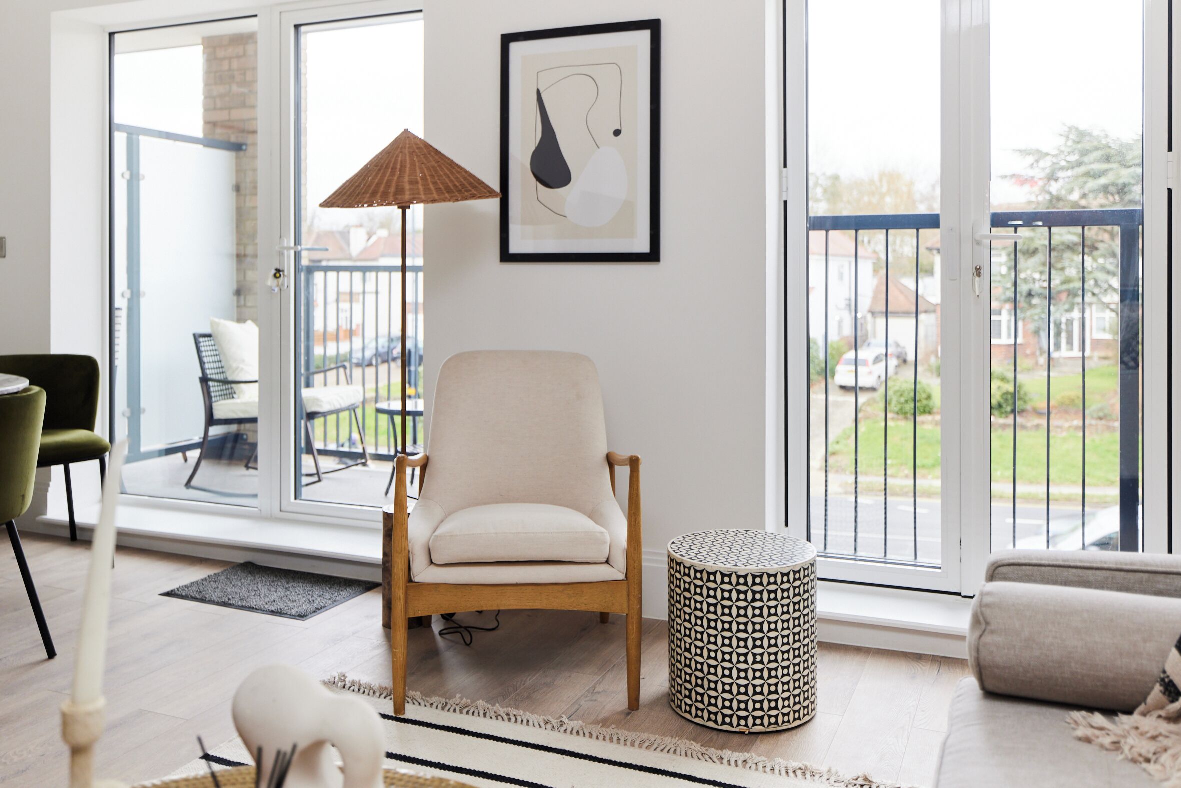 A modern living room with a large sliding glass door leading to a balcony, a beige armchair, a patterned ottoman, and a framed abstract artwork on the wall.