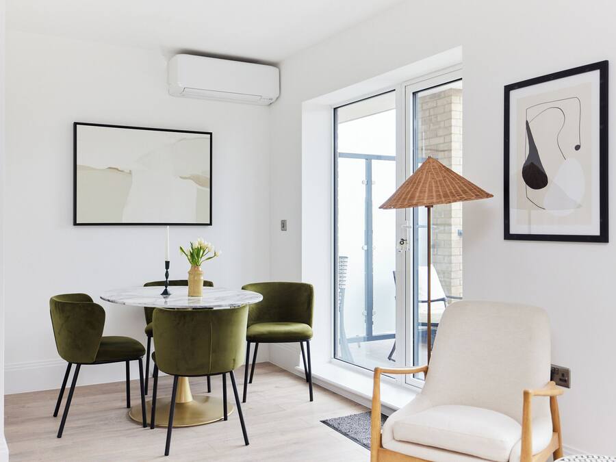 Un comedor moderno con una mesa redonda, dos sillas verdes y una silla blanca. Hay dos obras de arte enmarcadas en la pared, una ventana con vistas y una lámpara de pie.