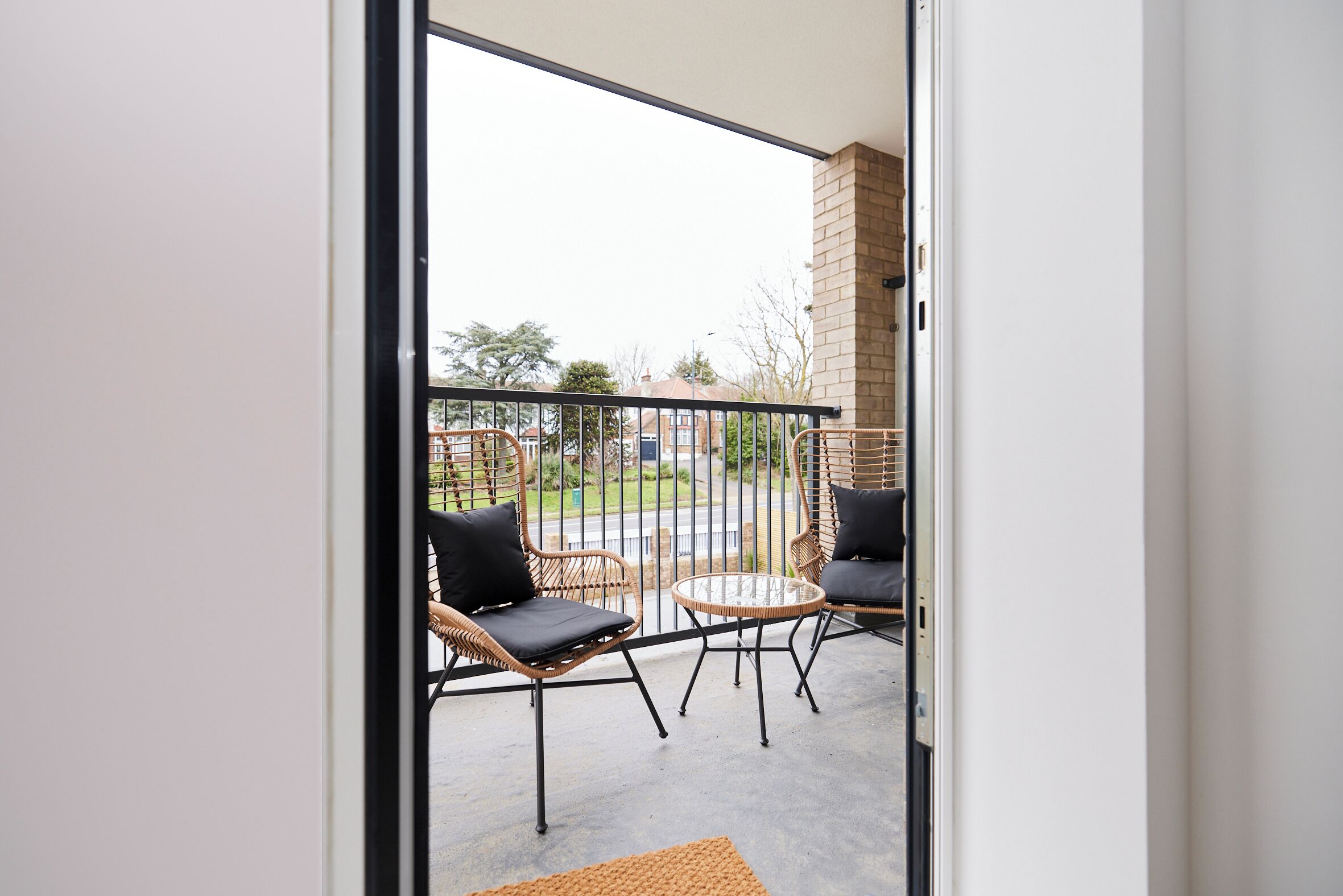 Un balcon avec des fauteuils en osier et une petite table, un mur en briques et une vue sur une rue et la verdure.