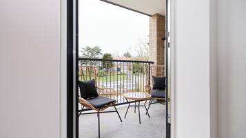Ein Balkon mit Korbstühlen und einem kleinen Tisch, eine Backsteinmauer und Blick auf eine Straße sowie Grünflächen.
