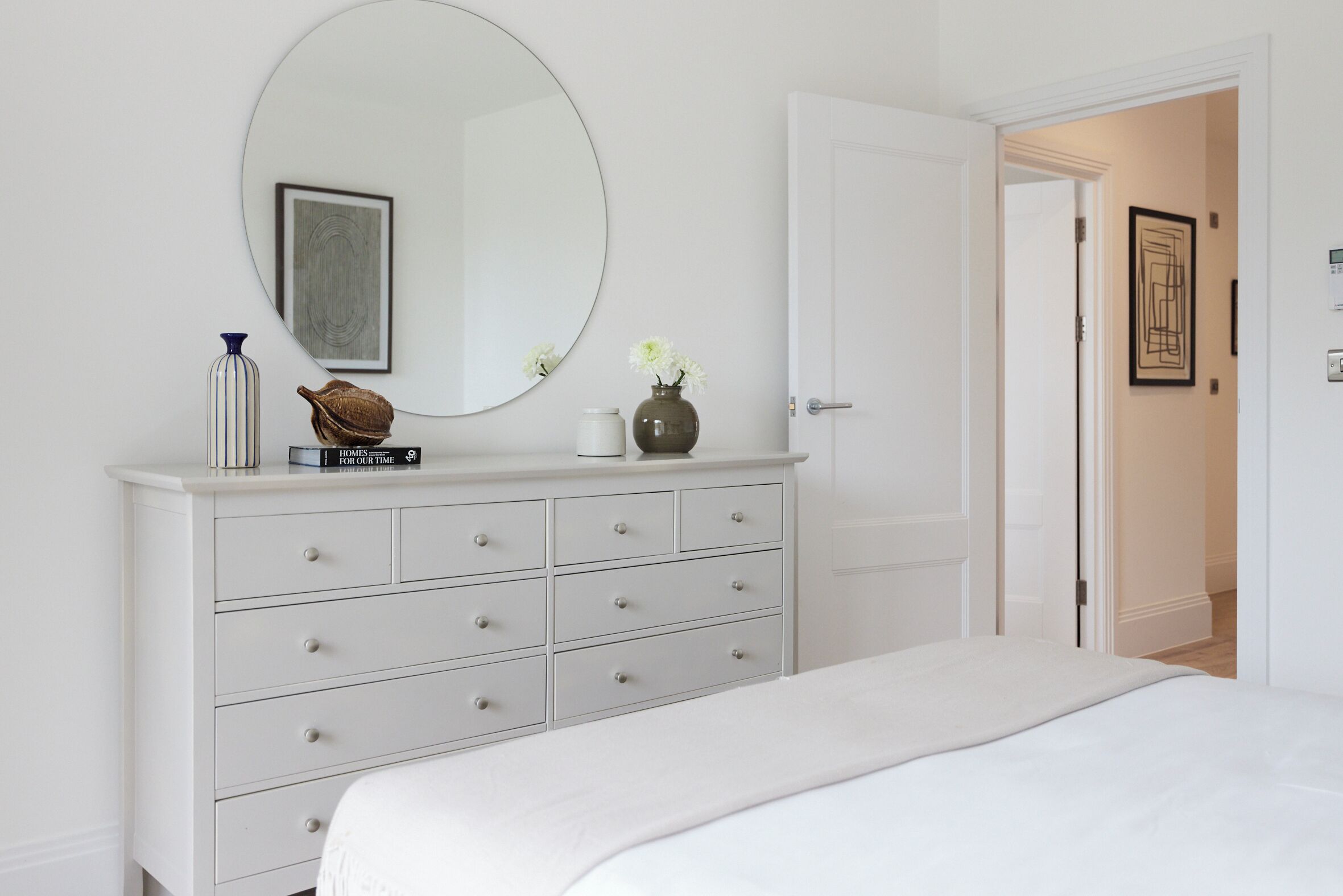 A bedroom with a white dresser, a round mirror, and a bed with white bedding.