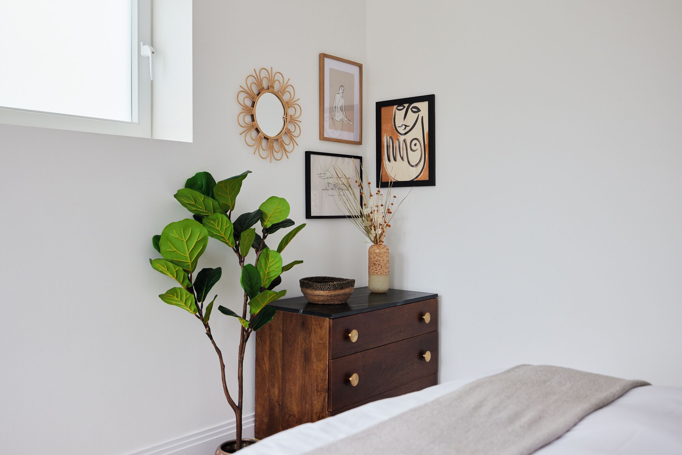 Une chambre à coucher avec un lit, une table de chevet dotée de tiroirs, une plante en pot et des œuvres d’art murales.