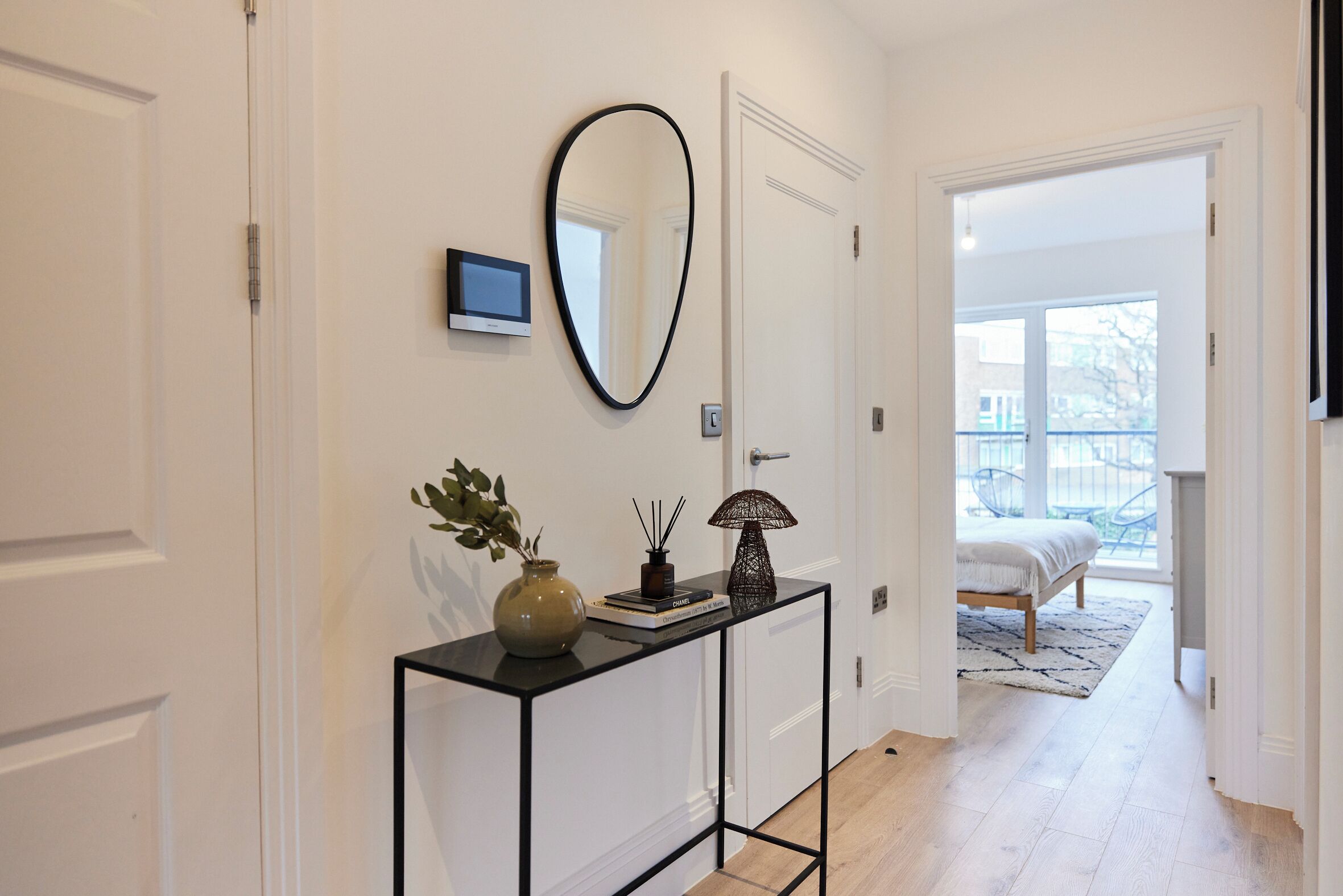 A hallway with a console table, a mirror, and a door leading to a bedroom with a bed and a view of the outside.