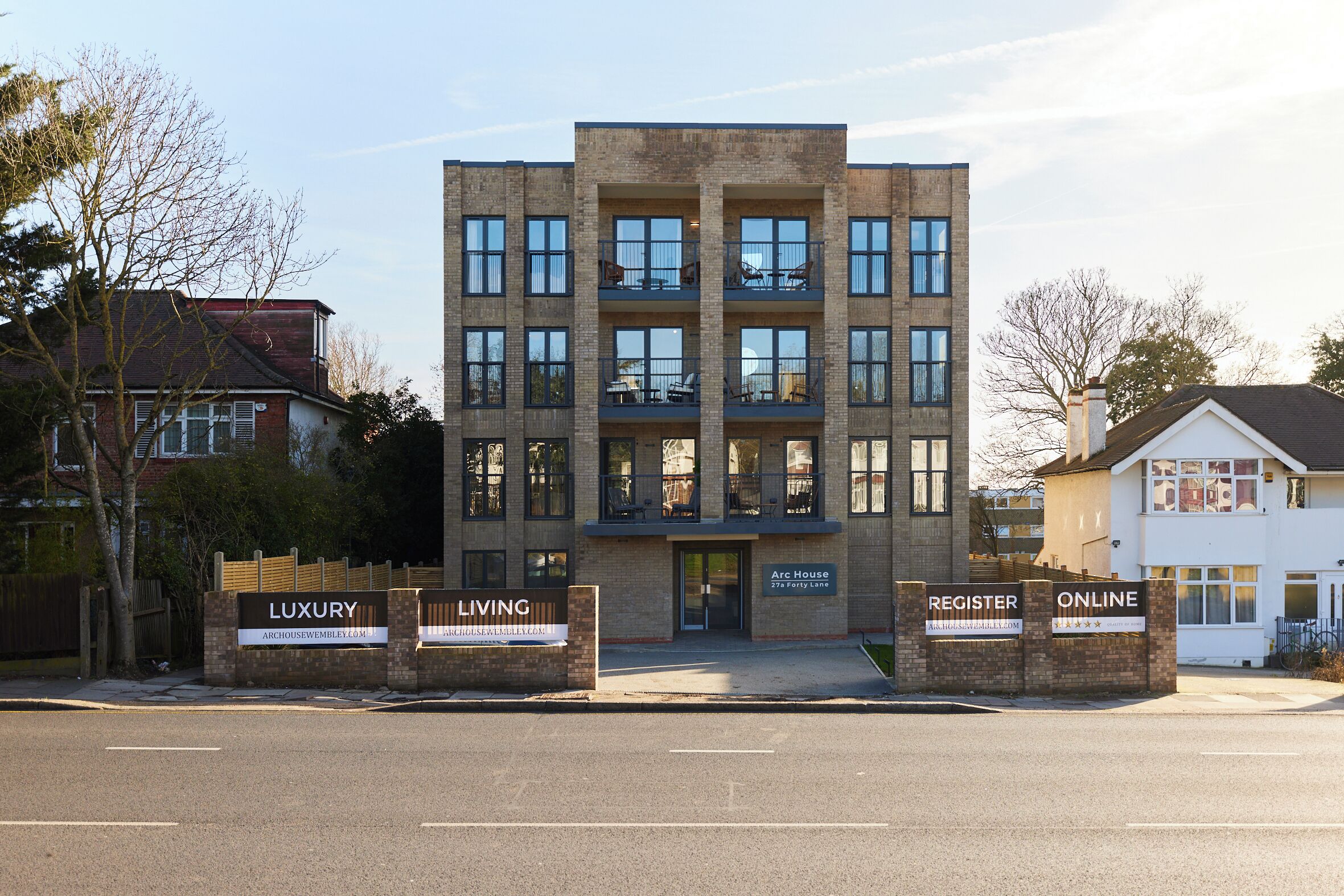 Un edificio moderno de varios pisos con fachada de ladrillo, ventanas grandes y balcones. Se observan letreros que indican 'Luxury Living' y 'Register Online'.