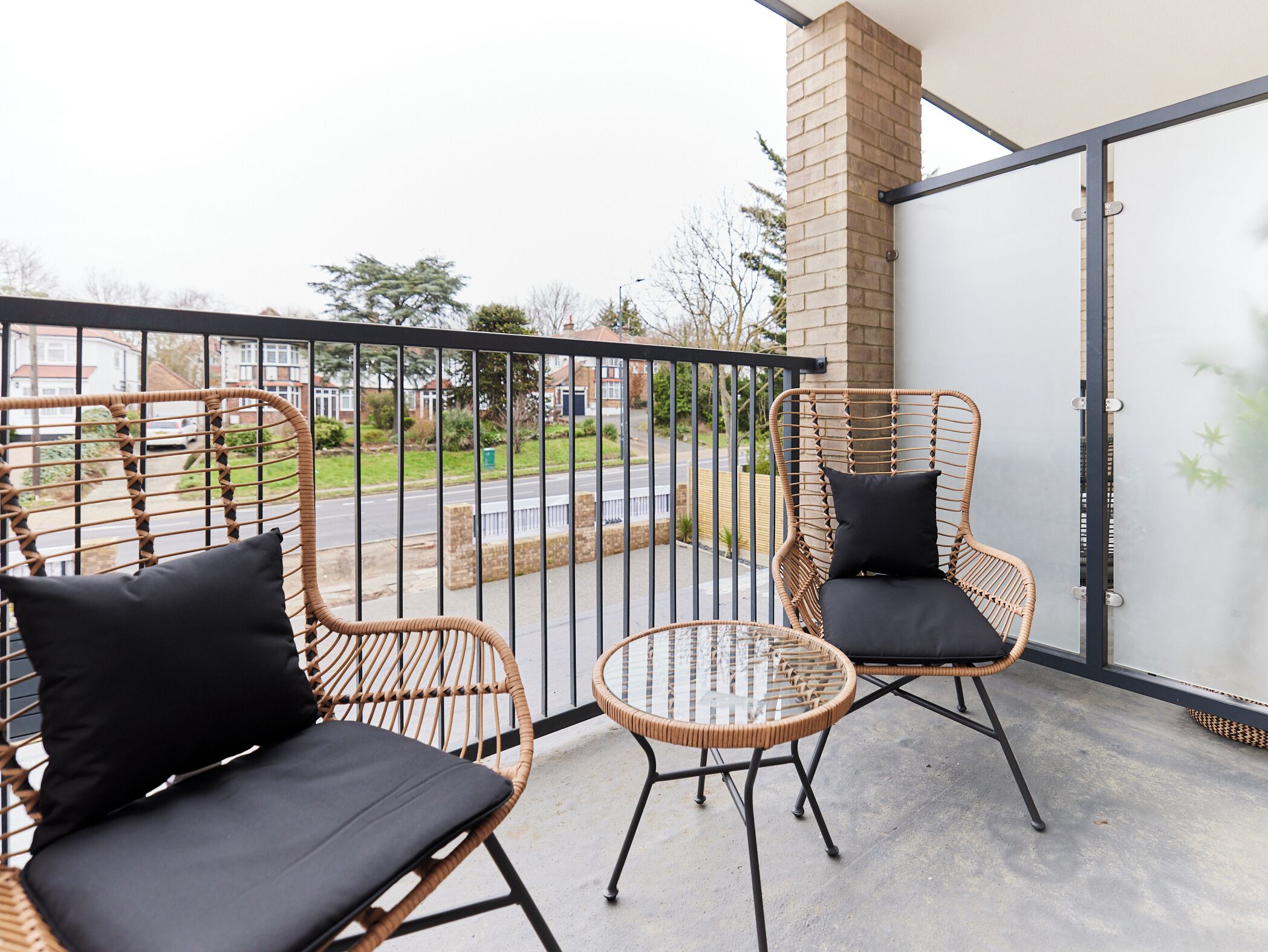 A balcony with wicker chairs and a glass table, overlooking a street and greenery.