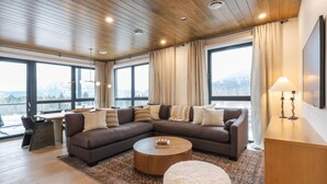 A modern living room with a sectional sofa, a round coffee table, and a view of the outdoors through large windows. - Mountain Magic at Mackenzie Village (Revelstoke)
