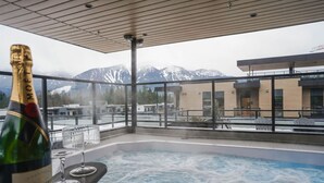 A hot tub with a mountain view, a bottle of champagne, and a modern building in the background. - Mountain Magic at Mackenzie Village (Revelstoke)
