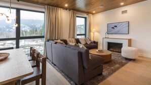 A modern living room with a sofa, a fireplace, and a large window offering a view of the outdoors. - Mountain Magic at Mackenzie Village (Revelstoke)