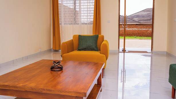 A room with a wooden table, a yellow armchair with a green pillow, and a view through a glass door to an outdoor area.