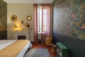 Ein Schlafzimmer mit einem Bett, einem Kamin, einem Fenster mit Vorhängen und einer Wand mit einem Blumenmuster.