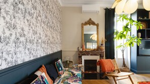 A room with a patterned wallpaper, a sofa with colorful cushions, a mirror, a fireplace, a chair, and a potted plant. - Charming Apartment in the Heart of Ajaccio (Ajaccio)