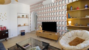 Een moderne woonkamer met een flatscreen televisie, een glazen salontafel, een rieten fauteuil en een behang met een patroon.