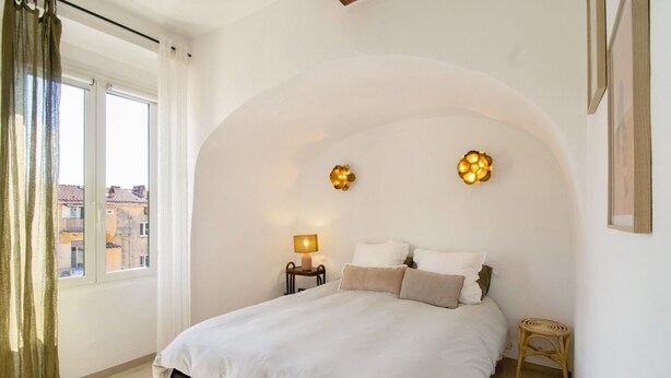 Een slaapkamer met een bed, een nachtkastje, een lamp, een klein krukje en een raam met gordijnen.