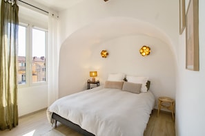 Un dormitorio con cama, mesita de noche, lámpara, taburete pequeño y ventana con cortinas.