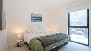 Ein Schlafzimmer mit großem Fenster, einem Bett mit weißer Bettwäsche, einer grünen Tagesdecke und einem kleinen Nachttisch mit Lampe.