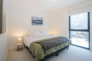A bedroom with a large window, a bed with white bedding, a green throw blanket, and a small bedside table with a lamp. - Zen Revy (Revelstoke)