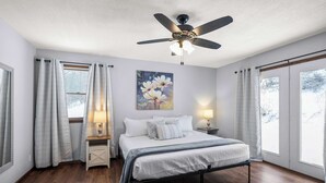 Une chambre à coucher avec un lit, deux tables de chevet avec des lampes, un ventilateur de plafond et de grandes fenêtres avec des rideaux.