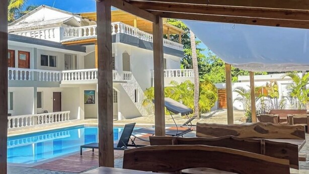 Un área de estar al aire libre cubierta con mesas y sillas de madera, con vista a una piscina y un edificio de varios pisos con balcones.