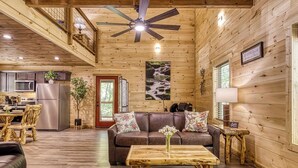 Interior rústico de una cabaña con techo de madera, una zona de comedor con mesa de madera, un sofá con cojines estampados y una cocina con refrigerador y microondas.