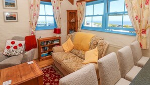 A cozy living room with a sofa, a wooden coffee table, and a bookshelf. Large windows with floral curtains offer a view of the sea. - Casa Mia (Goodwick)