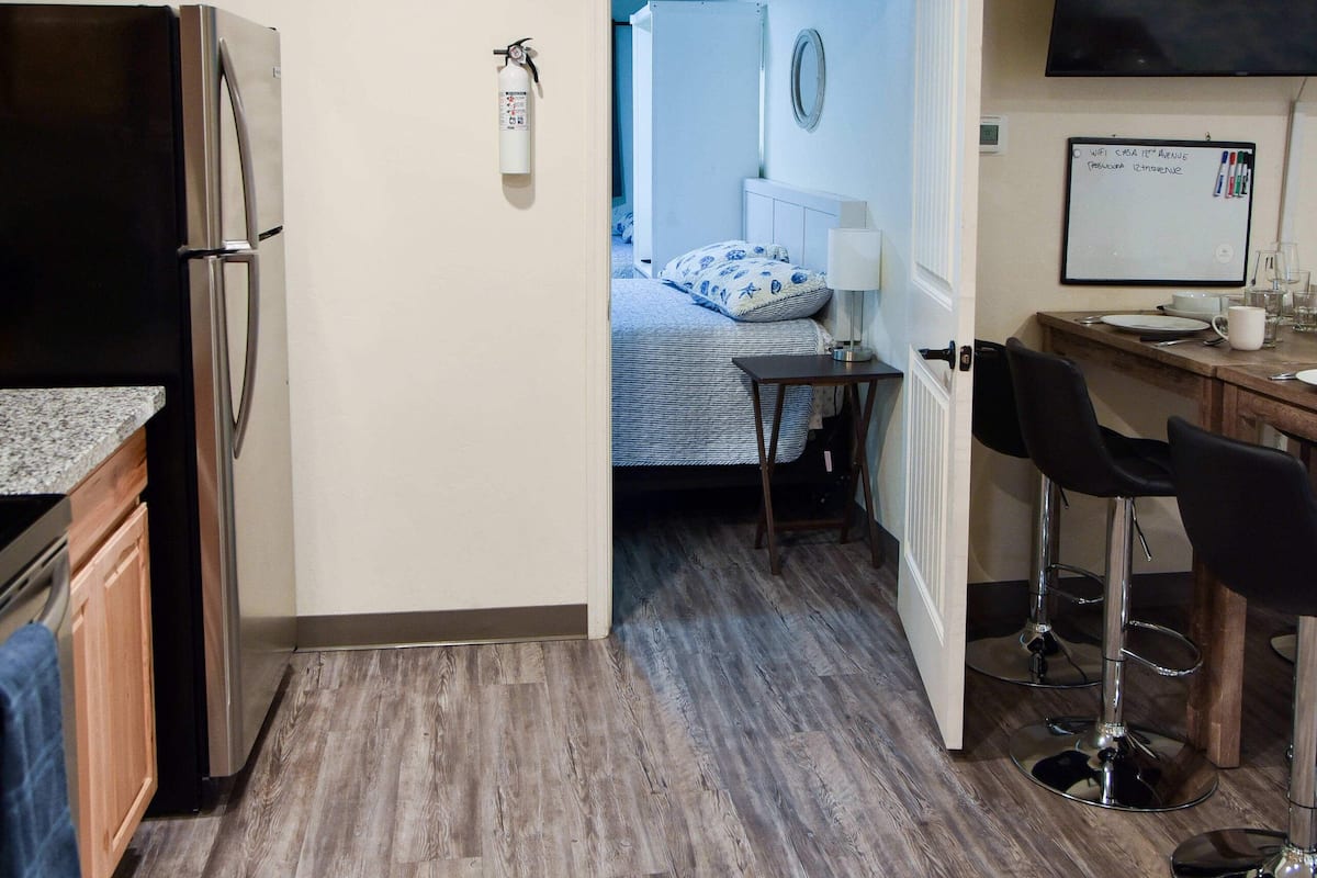 A compact hotel room with a kitchenette, a bed with a blue and white patterned bedspread, a small desk with a computer, and a TV mounted on the wall.