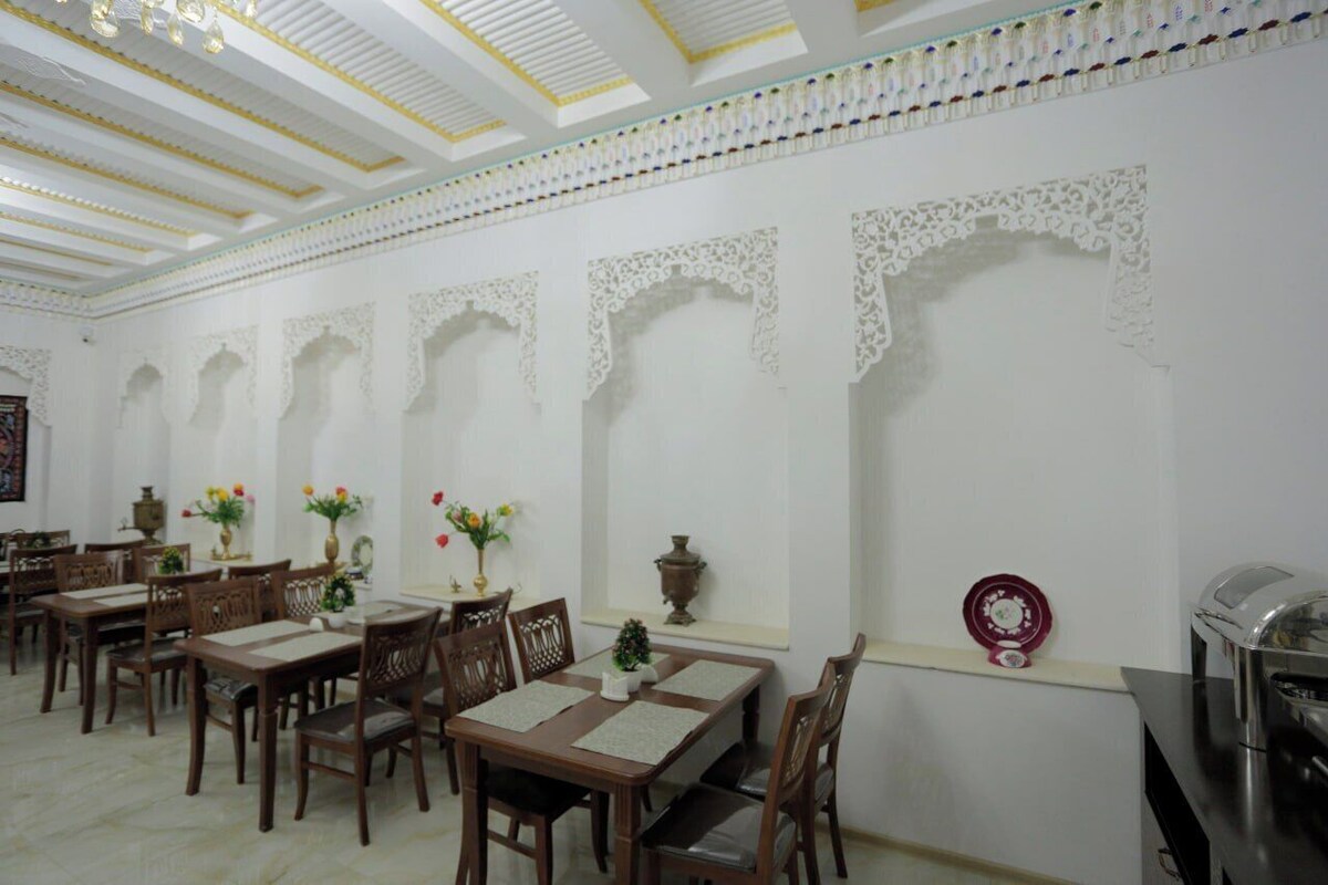 A dining area with wooden tables and chairs, white walls with decorative arches, and a ceiling with patterned beams.
