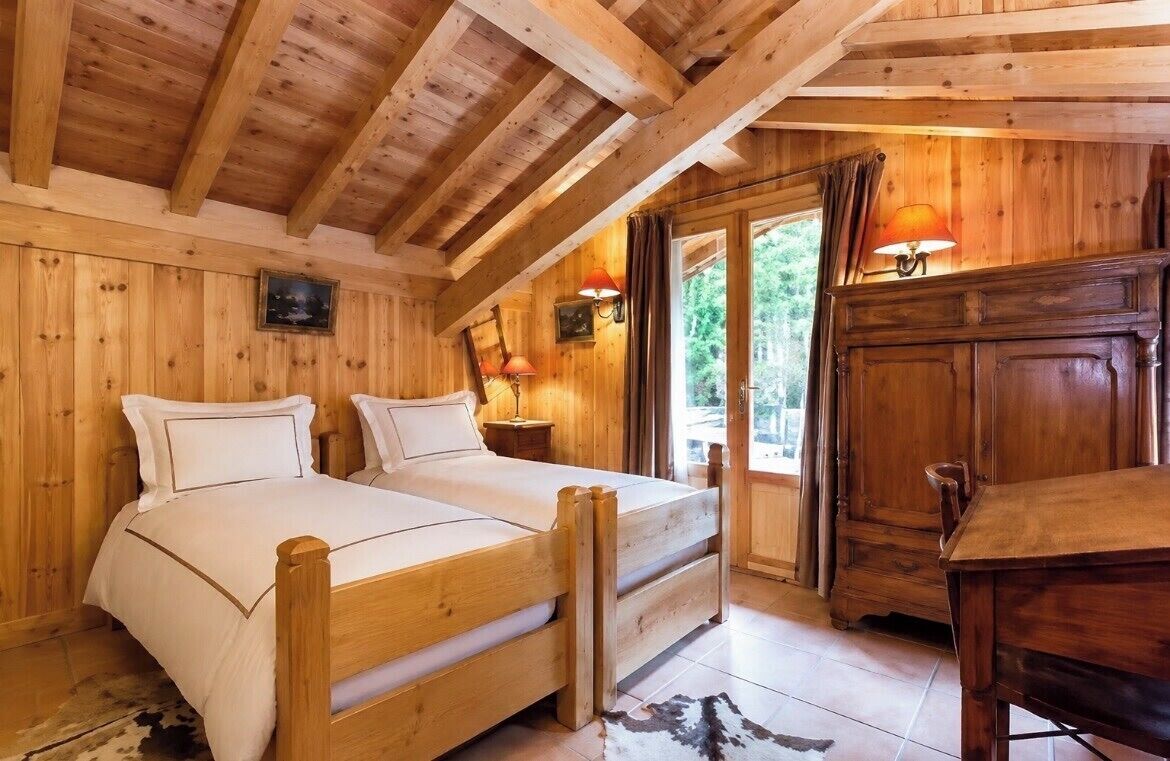 A wooden cabin bedroom with two beds, a nightstand, a dresser, and a window with curtains.