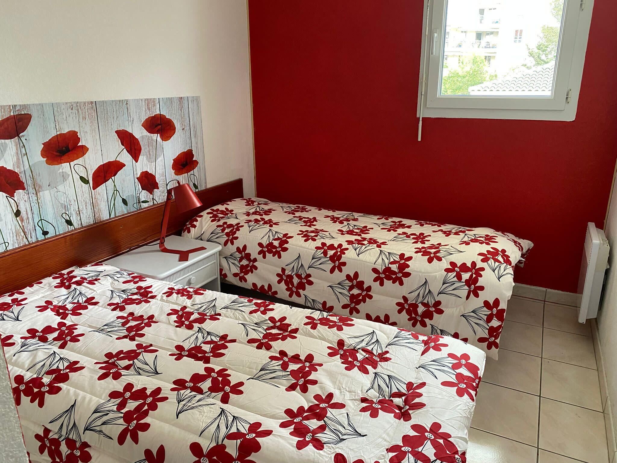 Une chambre avec deux lits, une tête de lit en bois, une chaise rouge et une fenêtre donnant sur des immeubles.