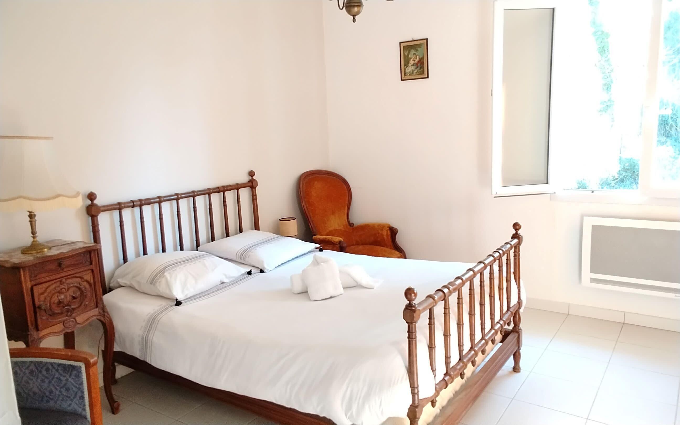 A neatly made bed with a wooden headboard, a nightstand with a lamp, a chair, and a window with a view of greenery.