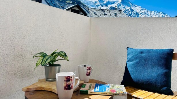 Een houten tafel met een plant, twee mokken, een boek en een kussen, met uitzicht op een berg tegen een muur.