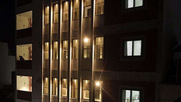 Ein mehrstöckiges Hotel mit beleuchteten Fenstern und Balkonen, ein sichtbares Schild mit der Aufschrift "AC ROOMS" und eine Straßenszene mit parkenden Fahrzeugen.