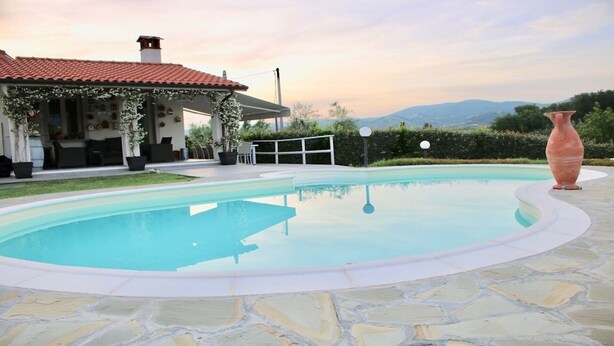 A swimming pool with a stone deck, a covered patio with outdoor furniture, and a view of mountains in the background. - Casa Il Cappero – Villa with Private Pool in the Tuscan Hills (San Rocco)