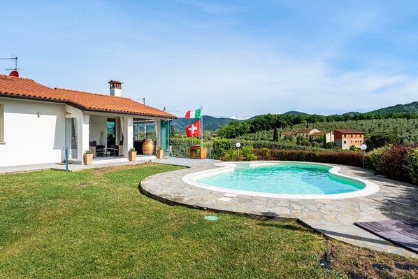 A pool area with a stone deck, a house with a red-tiled roof, and a flag.