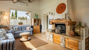 A cozy living room with a fireplace, stone fireplace surround, striped sofa, and a large window with a view of trees. - Casa Il Cappero – Villa with Private Pool in the Tuscan Hills (San Rocco)