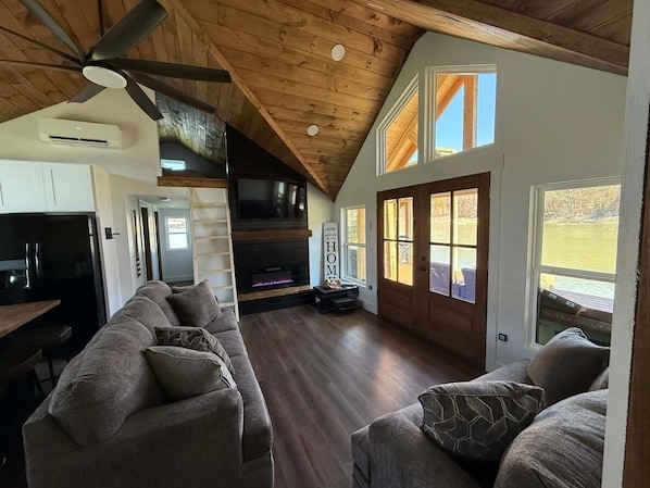 A spacious living area with a fireplace, wooden ceiling, and large windows.