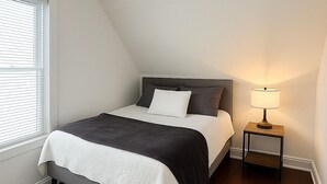 A bedroom with a bed, a bedside table with a lamp, and a window with blinds. -  Hidden Gem Steps From River, Restos, Cafés, Parks (Windsor)