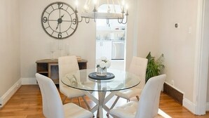 A dining area with a glass table surrounded by white chairs, a chandelier above, and a wall clock. -  Hidden Gem Steps From River, Restos, Cafés, Parks (Windsor)