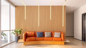 A modern lobby with a curved orange leather sofa, a wooden slatted wall, and a large potted plant. - Luxury studio apartment, do everything on foot! (São Caetano do Sul)
