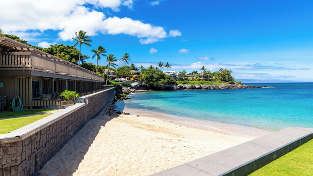 A beachfront property with a stone wall, a sandy beach, and a clear blue ocean. - Beachfront 2BR Townhouse w/ Ocean Sunset Views (Lahaina)