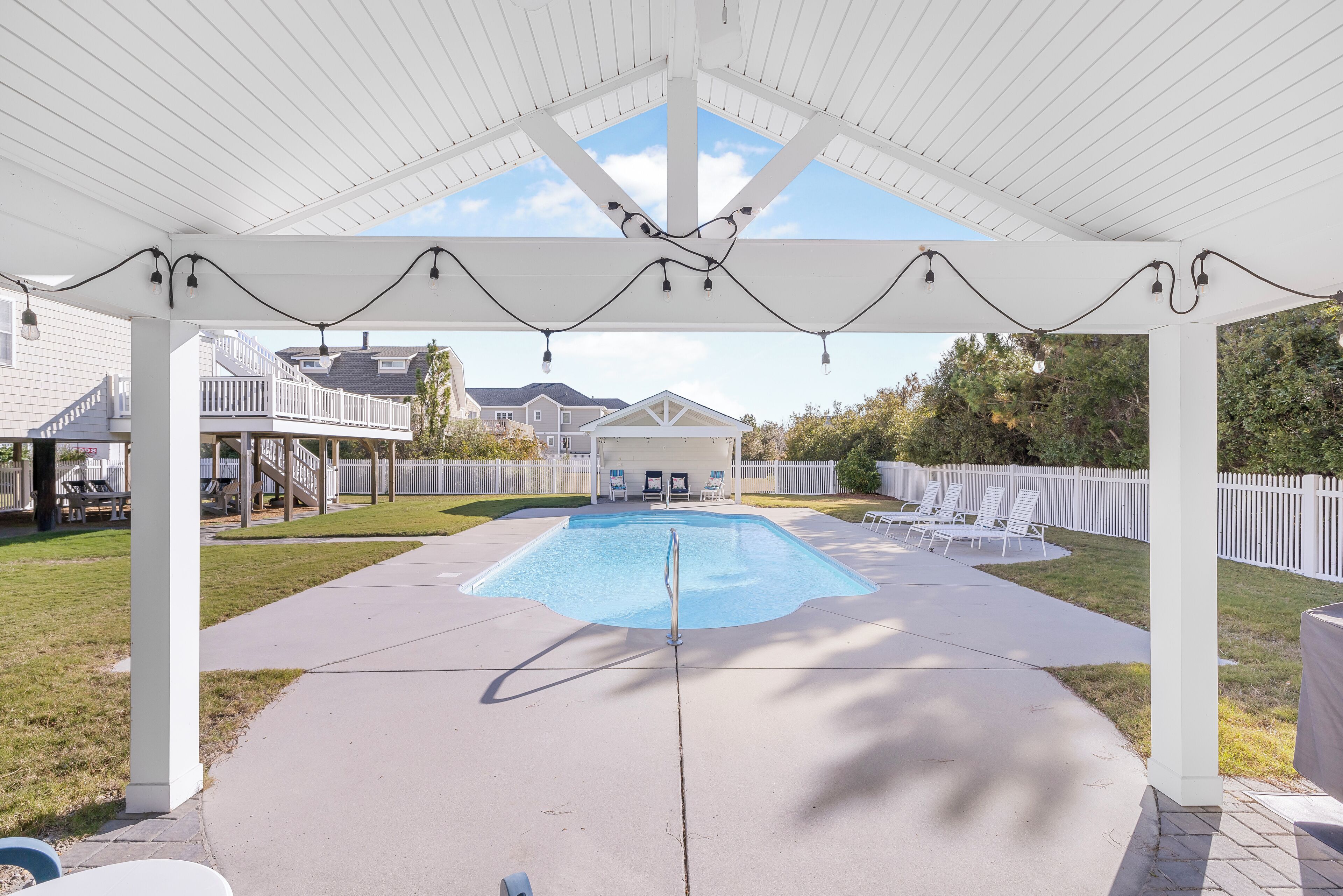 Una piscina cubierta al aire libre con un pabellón blanco, sillones de descanso y un pequeño edificio al fondo.