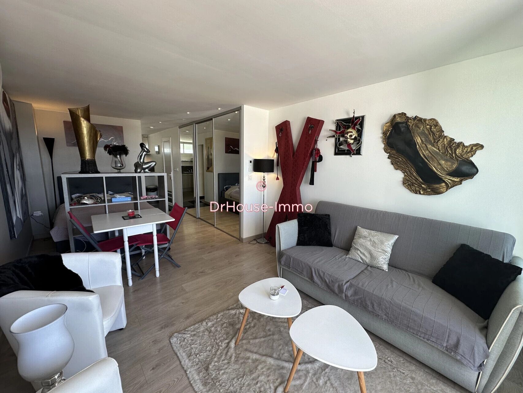 A modern living room with a grey sofa, white coffee table, and red chairs. There is a dining area with a white table and chairs. The room has a large mirror and decorative wall art.