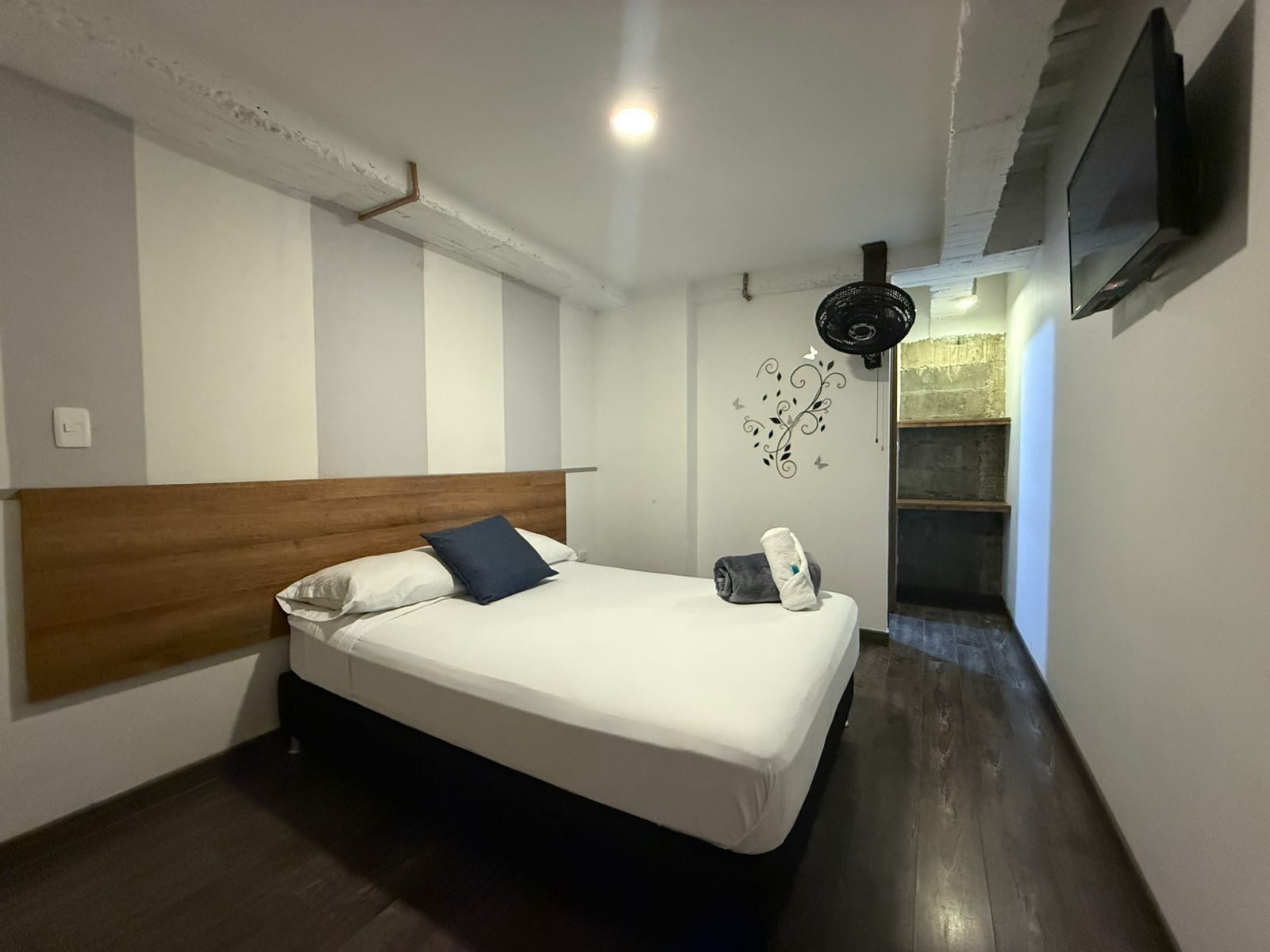 A bedroom with a wooden headboard, a bed with white bedding, a wall-mounted TV, and a ceiling fan.