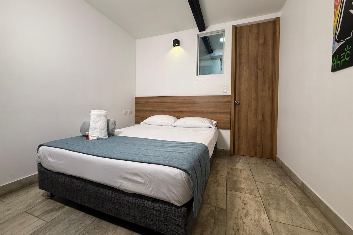 Un dormitorio con cama, cabecero de madera, una ventana pequeña, una puerta y una lámpara fijada a la pared.