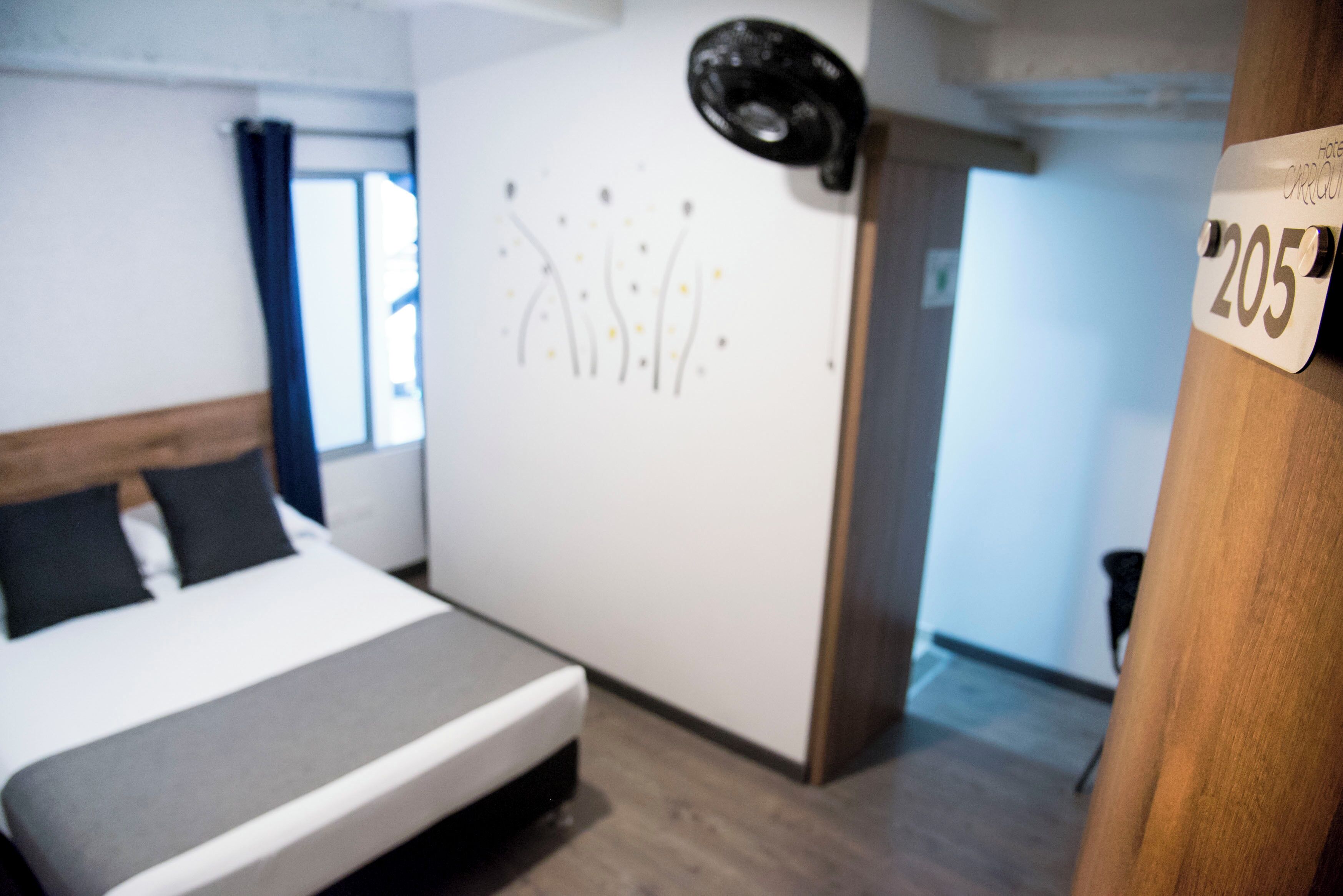 A modern hotel room with a bed, a wall-mounted light fixture, and a door with a number sign.