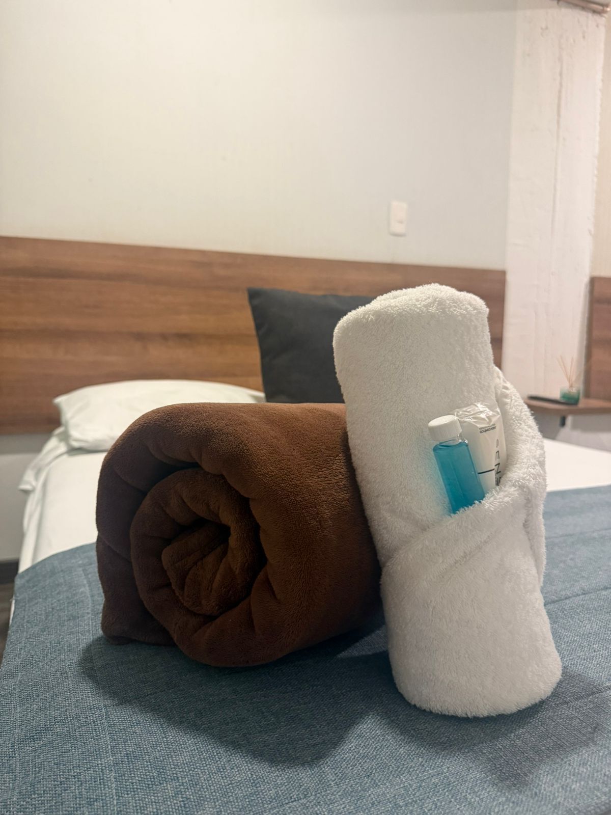 Una cama con una colcha azul, una toalla marrón y una toalla blanca junto a una botella.