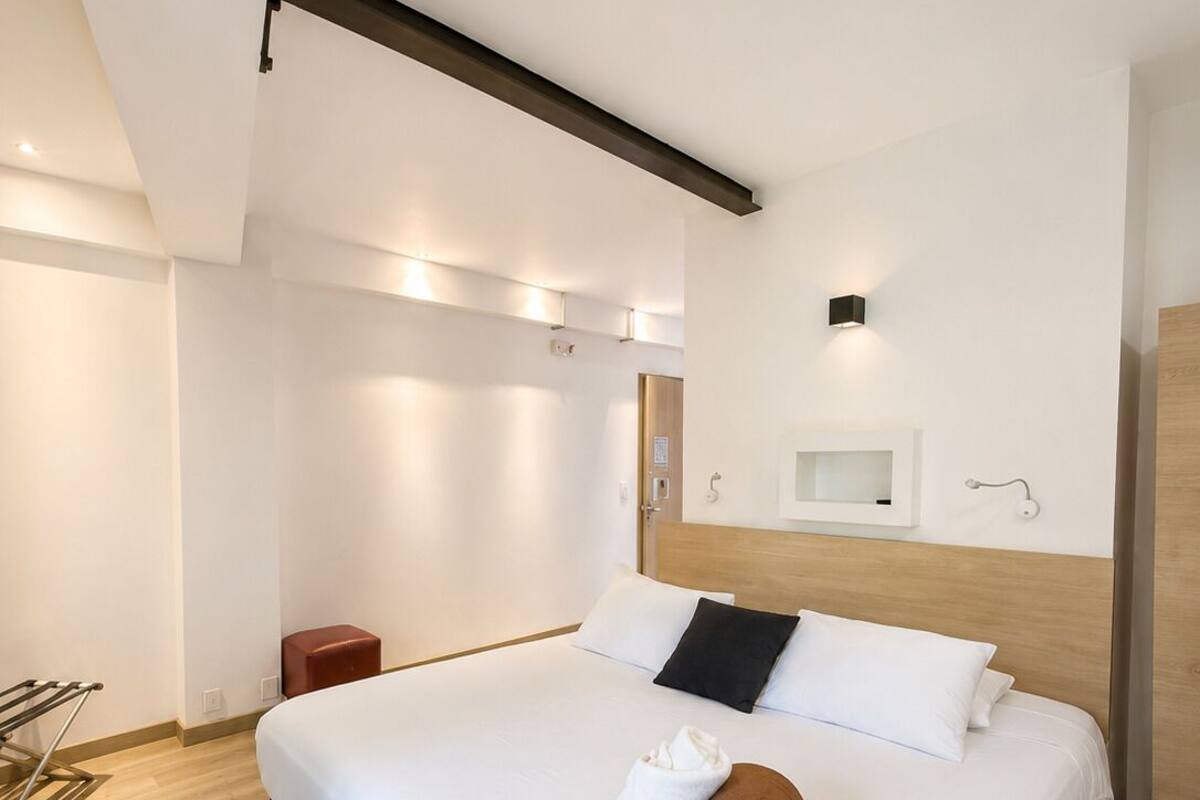 Una habitación de hotel moderna con una cama grande, un cabecero de madera, una mesita de noche pequeña y una estantería de pared.